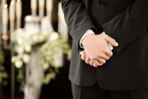 Image is of a closeup of a man's hands clasped together in front of him with funeral flowers in the background, concept of Melbourne wrongful death lawyer