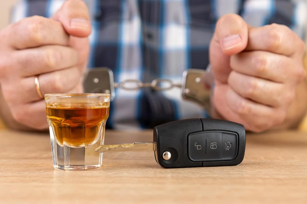Image is of a person in handcuffs with a drink and car key on the table, showing how legal trouble and alcohol use connect to the effect of a DUI on insurance.
