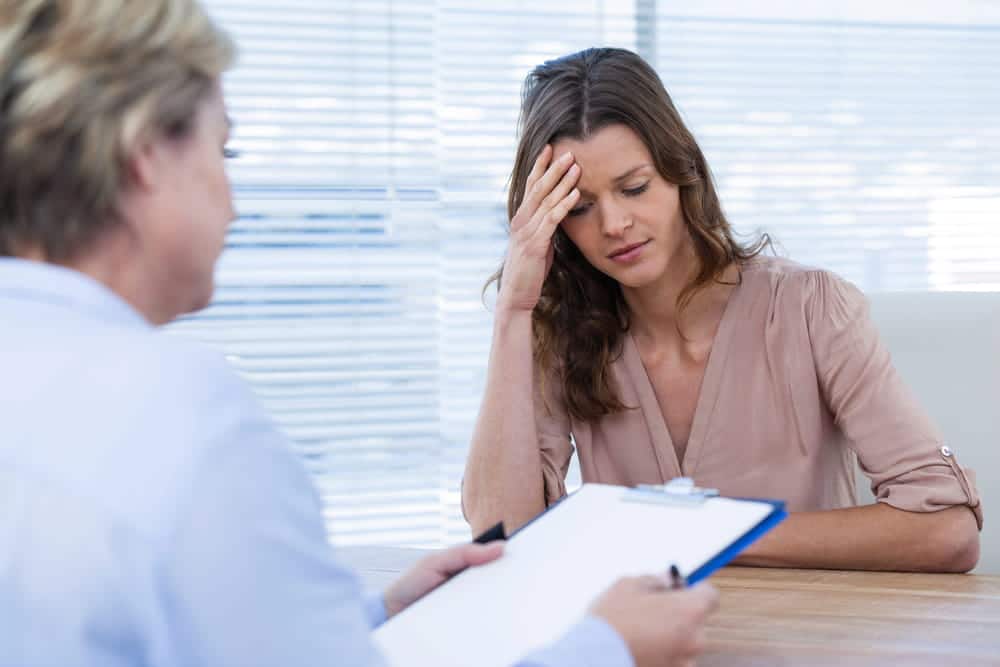 Image is of a woman speaking with a counselor while holding her head in concern, concept of domestic violence and mental health support and evaluation.
