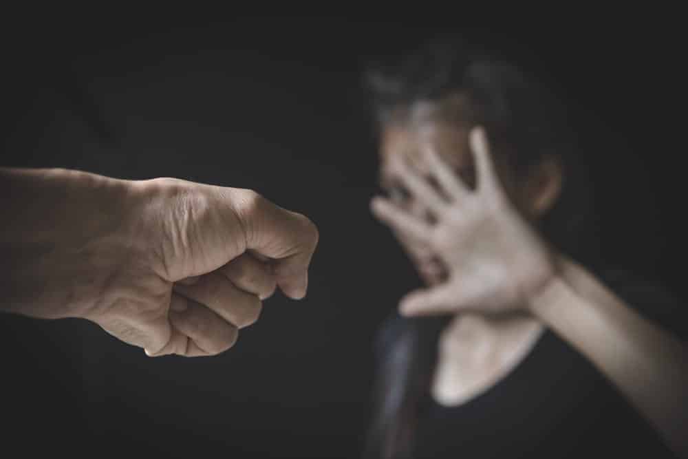 Image is of a raised fist near a distressed person in the background, concept of aggravated domestic violence involving threats and intimidation.