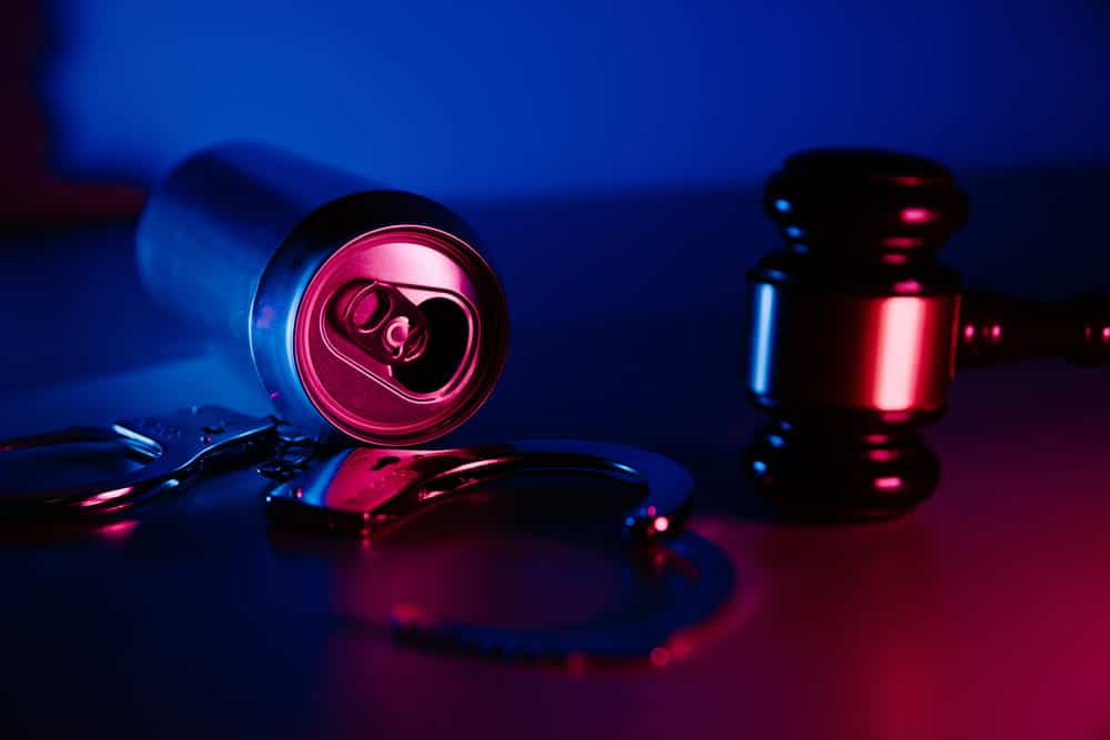 Image is of a judge's gavel, handcuffs, and an alcoholic beverage can on a table, concept of DUI penalties and criminal court proceedings.
