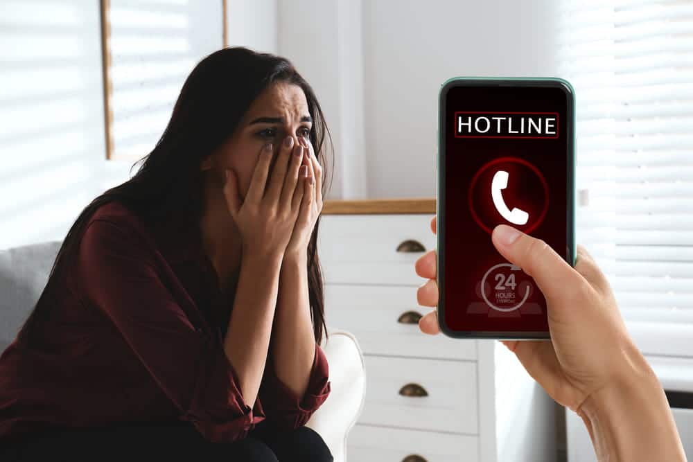 Image is of a distressed woman beside a phone displaying a hotline call, concept of a domestic violence victim seeking help.